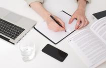 Close up of young man hands writing notes, making abstract, summary, opened a notebook on the table, there is a laptop near by, glass of water, phone with black screen, book and notebook