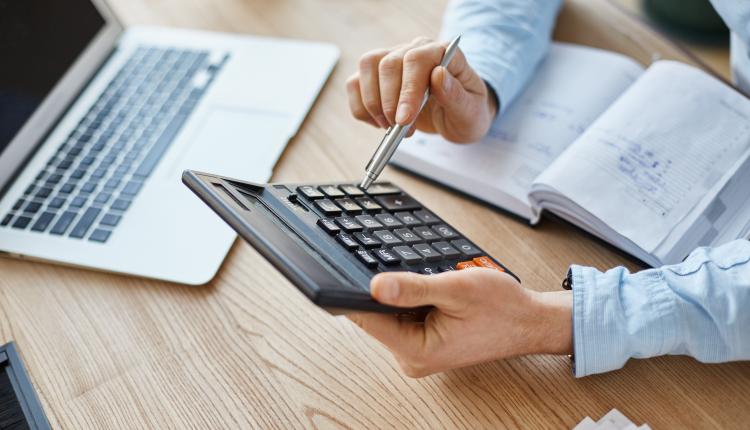 Cropped view of professional serious finance manager, holding calculator in hands, checking company month's profits, looking through details on laptop, writing down information for meeting with director.