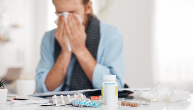 Portrait of ill, sick bearded male sneezes and coughs, uses handkerchief, rubs nose. Man has running nose, caugh, bad cold, sits at work place, surrounded by pills, drugs, and vitamins.