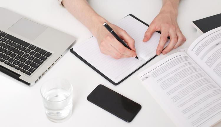 Close up of young man hands writing notes, making abstract, summary, opened a notebook on the table, there is a laptop near by, glass of water, phone with black screen, book and notebook