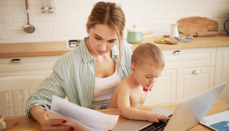 Serious concentrated young female studying papers in hands, paying bills online, sitting at kitchen table in front of open laptop holding baby son on her lap. Little child typing on portable computer